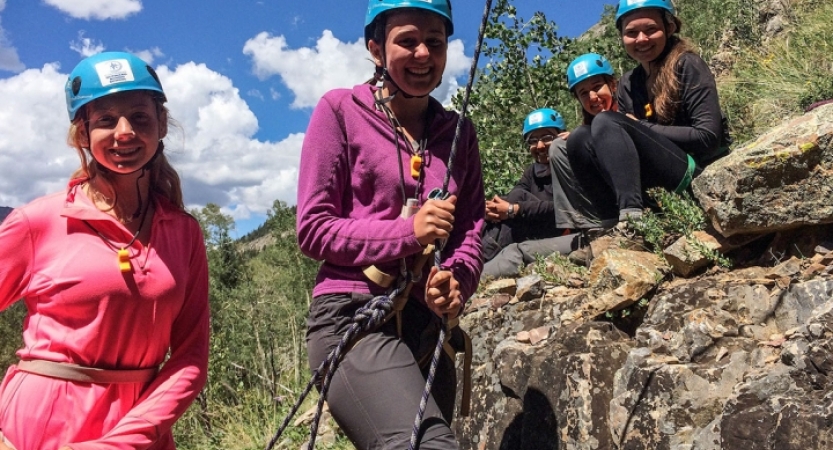 Outward bound students wearing helmets smile at the camera. One person appears to be belaying.